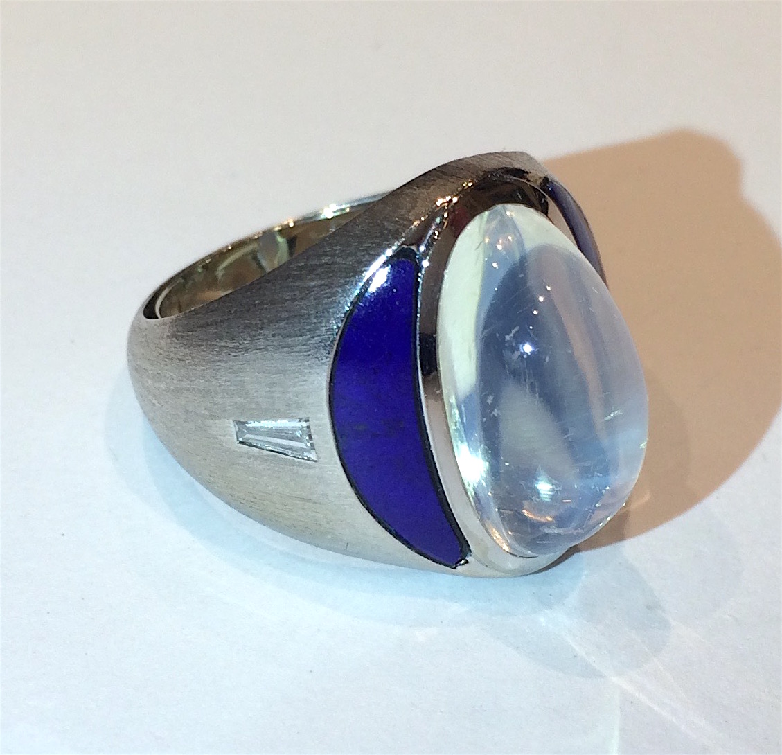 Guebelin Switzerland “Half moon” ring in 18K white brushed gold set with a large cabochon oval moonstone, two crescent moon shaped inset lapis lazuli stones and further set with two tapered baguette diamonds on either side, signed with the Guebelin cipher, 750 and Swiss, c. 1950’s