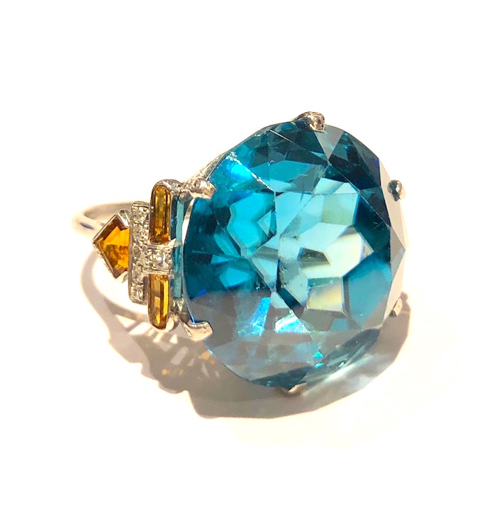 American Art Deco ring (likely by Louis C. Tiffany for Tiffany & Co.) set with a large blue zircon (approx. 64 carats by calculation, 20mm x 20mm x 19mm) set in a fancy platinum mount further set with 14 round cut diamonds, four bullet shaped gold/orange citrines and two kite shaped gold/orange citrines, marked: very deeply stamped letters as a maker’s mark on the inside of the shank, c. 1930